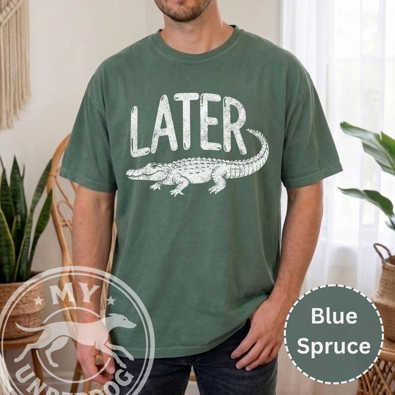 Pode incluir: Camiseta azul abeto com a palavra "LATER" acima de um gr&aacute;fico de jacar&eacute; branco. A camiseta de manga curta e gola redonda tem uma cor s&oacute;lida com um gr&aacute;fico desgastado. Estilo casual.