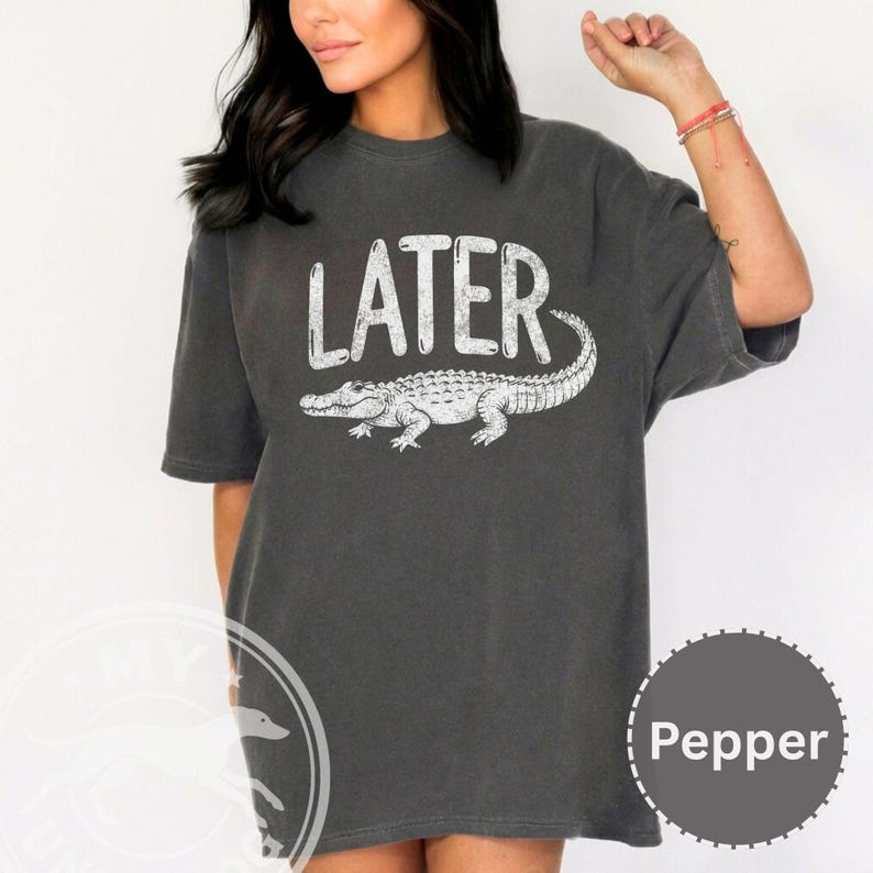 Pode incluir: T-shirt cinza escuro com a palavra "LATER" em branco, acima de uma ilustra&ccedil;&atilde;o de um jacar&eacute; branco. A camisa tem um ajuste folgado, com mangas curtas. A camisa &eacute; de cor s&oacute;lida, com decote redondo.