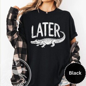Pode incluir: Camiseta preta com a palavra "LATER" em branco, acima de uma ilustra&ccedil;&atilde;o de um jacar&eacute; branco. A camiseta &eacute; de estilo casual, adequada para o uso di&aacute;rio. O design &eacute; simples e arrojado.