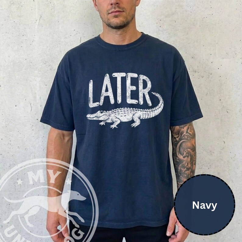 Pode incluir: Camiseta azul marinho com a palavra "LATER" em branco acima de uma ilustra&ccedil;&atilde;o de um jacar&eacute; branco. A camiseta de manga curta e gola redonda &eacute; de estilo casual.