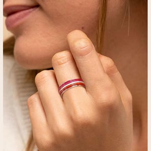 Puede incluir: Primer plano de una mano que lleva un anillo con tres bandas de color: rojo, blanco y rosa. El anillo está en el dedo anular. El fondo está borroso, con la cara y el hombro de una persona visibles.