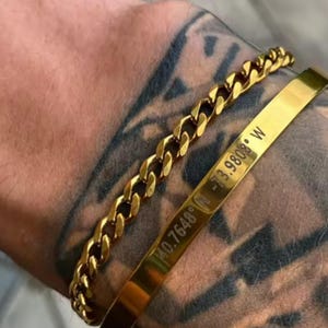 Op de afbeelding: Twee gouden armbanden op een getatoeëerde arm. Eén is een gourmetteketting, de andere is een massieve band gegraveerd met coördinaten: 40.7648° N -73.9808° W. De armbanden hebben een warme, glanzende goudkleur.