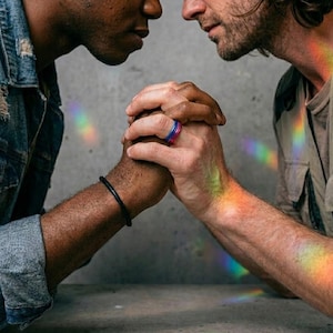 May include: Two people holding hands, with a rainbow light effect across their arms and faces. One person wears a black bracelet. One person wears a ring with blue, pink, and purple bands. The background is a gray concrete wall.
