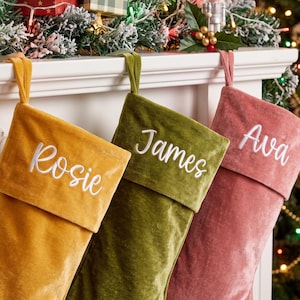 Op de afbeelding: Drie kerstsokken van fluweel in mosterdgeel, olijfgroen en oudroze. Elke sok heeft een omgeslagen boord met een naam geborduurd in wit schrift: Rosie op de gele, James op de groene en Ava op de roze kerstsok.