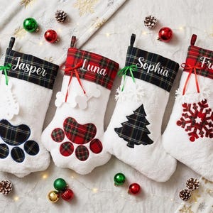 May include: Four white plush Christmas stockings with personalized names Jasper, Luna, Sophia, and Frank. The stockings feature plaid cuffs and decorative accents including paw prints, a Christmas tree, and a snowflake, tied with ribbons.