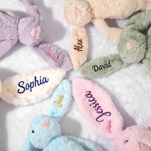 May include: A collection of plush bunny toys in various pastel colors, including lavender, cream, and blue. Each bunny has an ear embroidered with a name: Grace, Alex, David, Sophia, Jack, and Jessica. The bunnies are arranged on a soft, white surface.