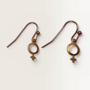 May include: A pair of bronze-toned metal drop earrings featuring a circular symbol with a downward-pointing cross. Each earring is attached to a curved ear wire hook with a small spherical bead at the end, creating a simple and uniform design.