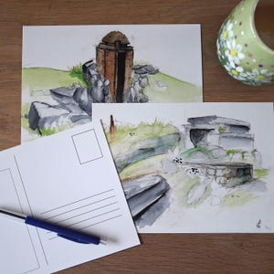 Op de afbeelding: Twee aquarelpostkaarten met architectonische schetsen van stenen structuren. Eén ansichtkaart toont een bruine, koepelvormige structuur met rotsen en groen gras. De andere toont een stenen gebouw met een plat dak. Een blauwe pen en een bloemenmok zijn ook aanwezig.