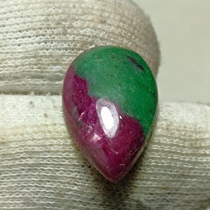Könnte beinhalten: Ein tropfenförmiger Edelstein mit polierter Oberfläche. Der Edelstein weist einen lebendigen Farbverlauf auf, der von einem tiefen Rot an der Basis zu einem satten Grün an der Spitze übergeht. Der Stein wird von einem weißen Handschuh gehalten.