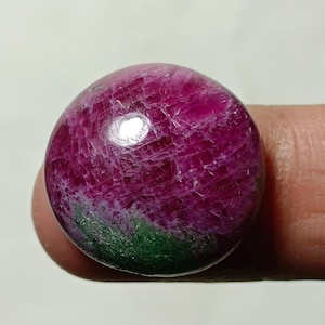 Könnte beinhalten: Ein polierter, runder Edelstein in tiefem Magenta, mit helleren Farbtönen geädert. Am Boden ist ein grünes Band zu sehen. Der Edelstein wird zwischen zwei Fingern gehalten.