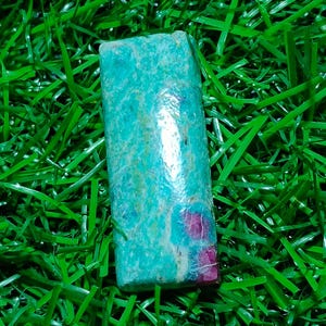 Könnte beinhalten: Ein rechteckiger Edelstein mit einem türkis-roten Muster. Der Stein ist vor einem Hintergrund aus grünem Kunstrasen platziert. Der Edelstein hat eine polierte, reflektierende Oberfläche.