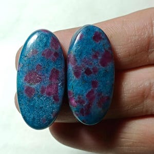Könnte beinhalten: Zwei ovale Edelsteine mit türkisfarbener Grundfarbe und tiefroten Flecken. Die polierten Steine haben eine glatte Oberfläche und zeigen ein natürliches, gesprenkeltes Muster. Geeignet für Schmuckherstellung.