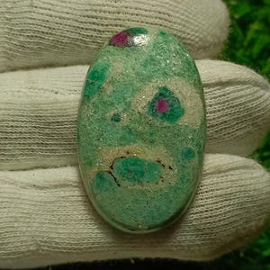 Könnte beinhalten: Ein ovaler Edelstein mit hellgrüner Grundfarbe, gesprenkelt mit dunklerem Grün und kleinen roten Einschlüssen. Der polierte Stein wird von einer behandschuhten Hand gehalten. Die einzigartige Farbe und das Muster des Edelsteins machen ihn zu einem Blickfang.