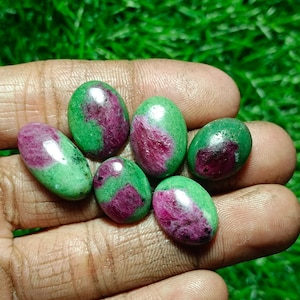 Könnte beinhalten: Eine Sammlung ovaler Edelsteine, die jeweils eine lebendige Kombination aus Grün und tiefroten Farbtönen aufweisen. Die polierten Steine werden in einer Hand gehalten, wodurch ihre glatten Oberflächen und natürlichen Farbvariationen hervorgehoben werden. Ideal für die Schmuckherstellung.