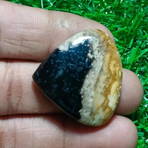 Puede incluir: Una piedra preciosa pulida con forma triangular, que presenta un llamativo contraste de colores. La piedra está dividida en tres secciones: un azul marino intenso, un blanco cremoso y una zona de color canela con motas marrones. La gema se sostiene en una mano.