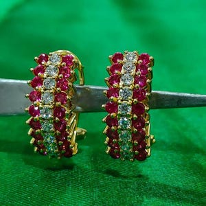 Puede incluir: Un par de pendientes de aro dorados. Cada pendiente presenta filas alternas de pequeñas piedras preciosas redondas de color rojo rubí y piedras transparentes de corte cuadrado. Los pendientes están sobre un fondo verde.