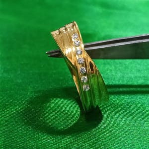 May include: A polished gold ring featuring a diagonal channel set with six small, clear, round-cut stones. The band has a smooth, reflective surface with subtle, raised parallel ridges that follow the curve of the metal across the ring's face.