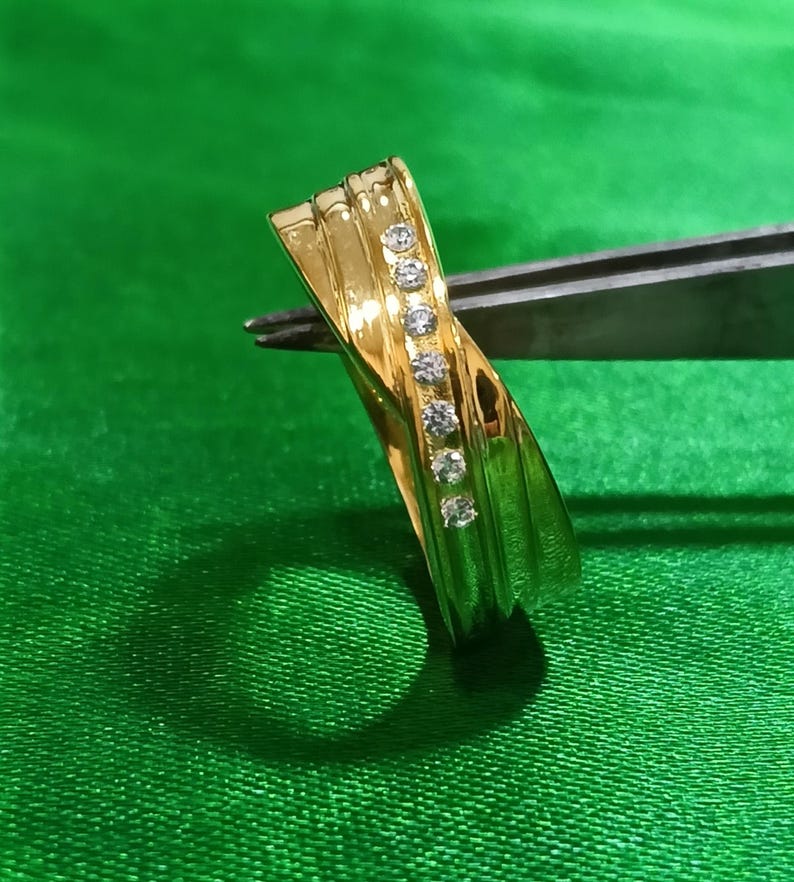 May include: A polished gold ring featuring a diagonal channel set with six small, clear, round-cut stones. The band has a smooth, reflective surface with subtle, raised parallel ridges that follow the curve of the metal across the ring's face.