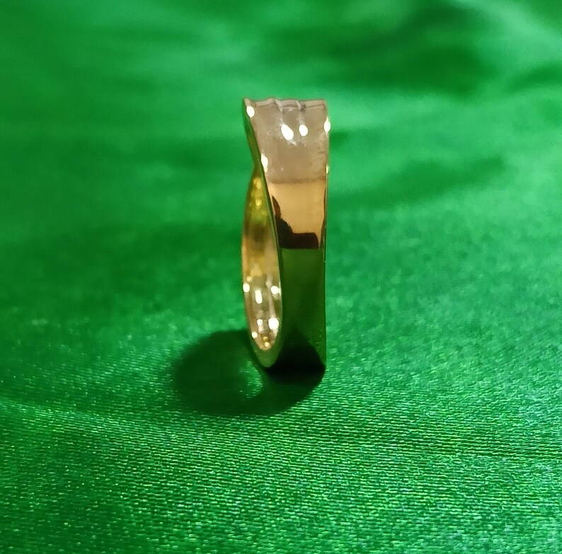 May include: A polished gold ring with a smooth, reflective surface and a slightly tapered, curved band design. The ring is positioned vertically, showcasing its metallic luster and rounded edges, reflecting light across its curved exterior.