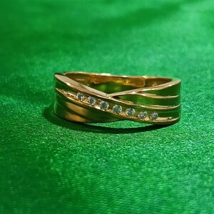 May include: A gold-toned band ring featuring a crossover design with a diagonal channel set with seven small, clear, round-cut stones. The metal surface is polished and reflective, creating a smooth, continuous loop with a tapered silhouette.