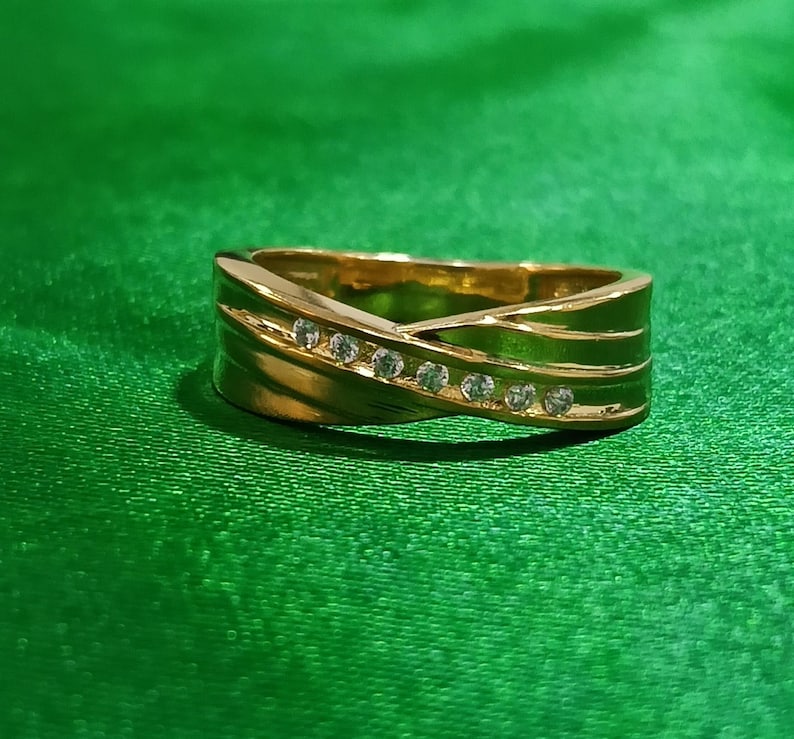 May include: A gold-toned band ring featuring a crossover design with a diagonal channel set with seven small, clear, round-cut stones. The metal surface is polished and reflective, creating a smooth, continuous loop with a tapered silhouette.