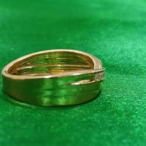 May include: A polished gold-tone ring featuring a wide, smooth band with a bypass-style design that overlaps at the front. A single, small, clear, round-cut stone is set into the side of the overlapping band, adding a subtle point of light.