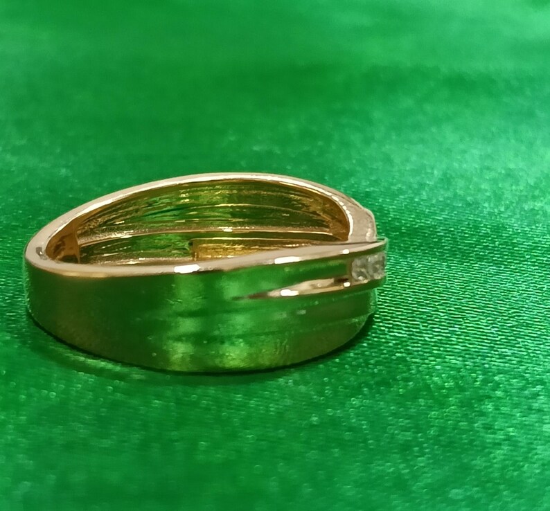 May include: A polished gold-tone ring featuring a wide, smooth band with a bypass-style design that overlaps at the front. A single, small, clear, round-cut stone is set into the side of the overlapping band, adding a subtle point of light.