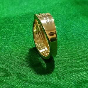 May include: A polished gold-tone metal ring featuring a smooth, wide band with a slightly raised, rounded center section. The surface of the ring is highly reflective, showing light highlights and a metallic finish throughout the entire piece.