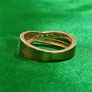 May include: A gold-toned ring featuring a crossover design with a smooth, polished band that tapers toward the center. The upper section of the band is accented with a row of small, clear, sparkling stones embedded along the overlapping metal curves.