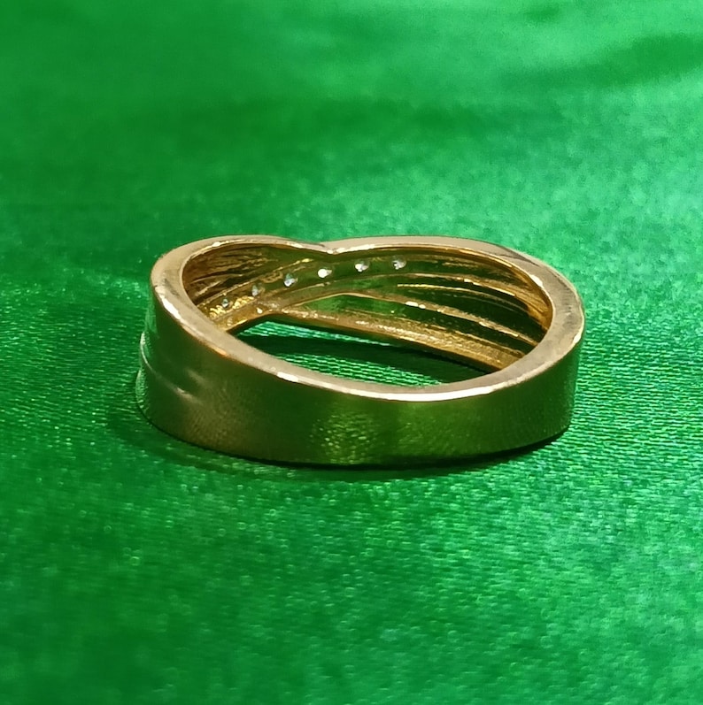 May include: A gold-toned ring featuring a crossover design with a smooth, polished band that tapers toward the center. The upper section of the band is accented with a row of small, clear, sparkling stones embedded along the overlapping metal curves.