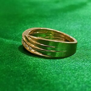 May include: A polished gold band ring featuring a smooth, wide surface with a diagonal groove detail. A small channel is set with three clear, round-cut stones that catch the light, creating a subtle sparkle against the metallic gold finish.