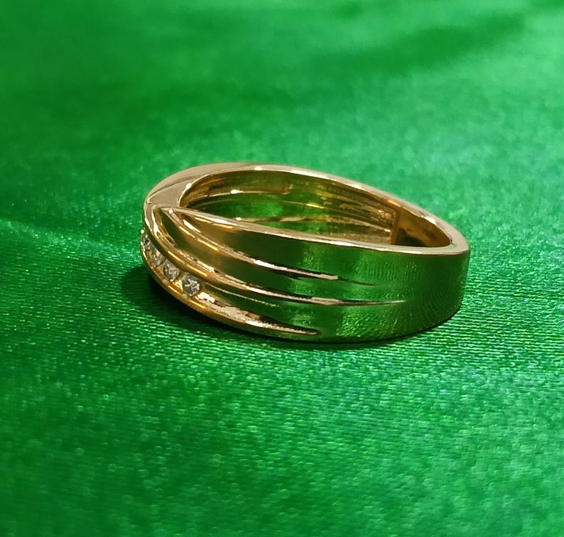 May include: A polished gold band ring featuring a smooth, wide surface with a diagonal groove detail. A small channel is set with three clear, round-cut stones that catch the light, creating a subtle sparkle against the metallic gold finish.
