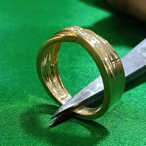 May include: A gold band ring featuring a polished finish and a central channel set with a row of small, clear, sparkling stones. The ring has a slightly twisted or crossover design element on the top surface, showcasing a smooth, reflective metallic texture.