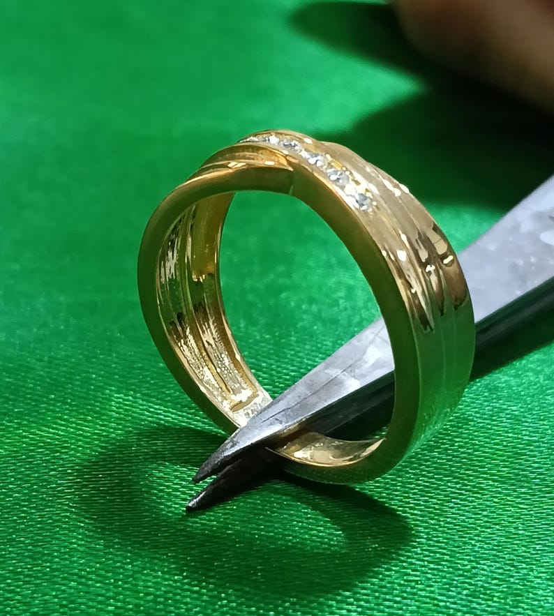 May include: A gold band ring featuring a polished finish and a central channel set with a row of small, clear, sparkling stones. The ring has a slightly twisted or crossover design element on the top surface, showcasing a smooth, reflective metallic texture.