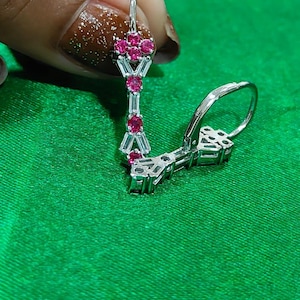 May include: Silver drop earrings with a geometric design, featuring clusters of small, round, bright pink gemstones and rectangular clear stones. The earrings are displayed against a green background.
