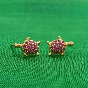 Könnte beinhalten: Ein Paar goldene Ohrstecker in Schildkrötenform. Jede Schildkröte hat einen Panzer, der mit kleinen, runden, leuchtend rosa Steinen besetzt ist, und einen winzigen klaren Stein im Kopf als Auge, inklusive eines Ohrsteckerverschlusses.