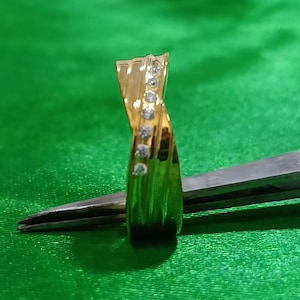 May include: A gold ring featuring a twisted band design with a vertical row of seven small, clear, round-cut stones embedded along the center. The polished metal surface reflects light, highlighting the curved structure of the band.