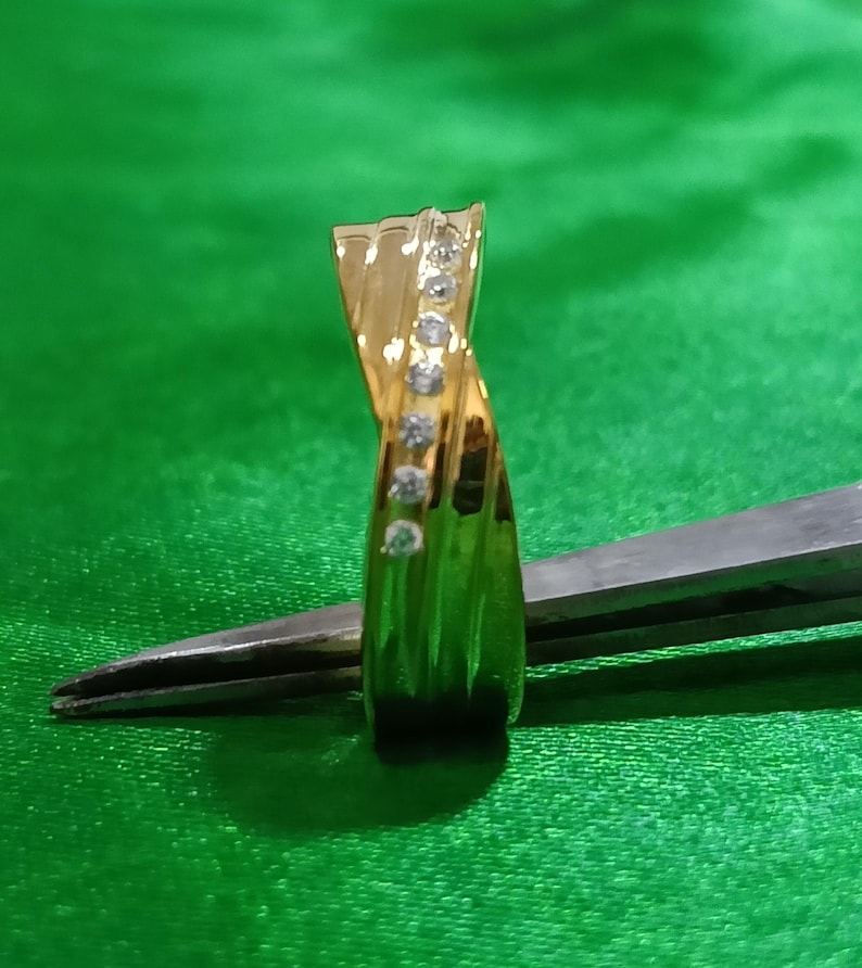May include: A gold ring featuring a twisted band design with a vertical row of seven small, clear, round-cut stones embedded along the center. The polished metal surface reflects light, highlighting the curved structure of the band.