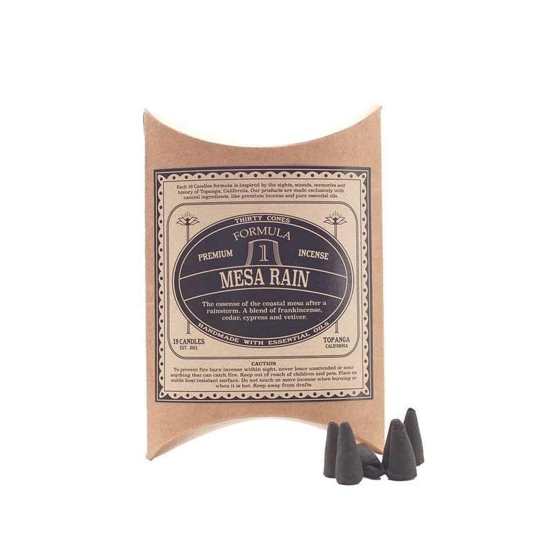 Puede incluir: Un paquete de conos de incienso "Mesa Rain" con un respaldo de papel marr&oacute;n. La etiqueta presenta texto y un dise&ntilde;o ovalado. Varios conos de incienso gris oscuro son visibles frente al paquete. La etiqueta indica que el incienso est&aacute; hecho a mano con aceites esenciales.