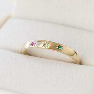 May include: A gold ring with three inlaid gemstones in a white jewellery box. The ring features a pink, white, and green gemstone. The ring is set against a white background.