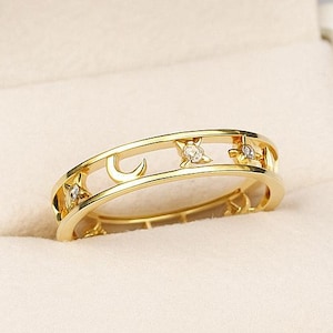 Puede incluir: Un anillo de oro con un diseño celestial, con estrellas, lunas y pequeñas piedras transparentes. El anillo se muestra dentro de una caja de joyería blanca con un interior de color crema.