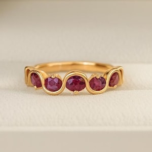 Może przedstawiać: Złoty pierścionek z serią owalnych, ciemnoczerwonych kamieni szlachetnych ułożonych w falisty wzór. Pierścionek jest prezentowany w białym pudełku na biżuterię, z miękkim, rozmytym tłem.
