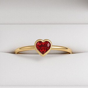 Puede incluir: Un anillo de oro con una piedra preciosa roja en forma de corazón, engastada en un bisel. El anillo se muestra dentro de una caja de joyería blanca, creando un contraste llamativo.