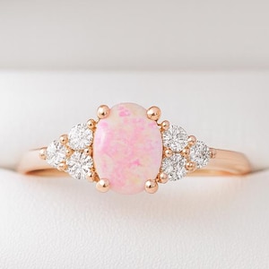 May include: A rose gold ring featuring a large oval pink opal center stone, flanked by clusters of smaller, clear gemstones. The ring is set in a white jewelry box, showcasing its elegant design. A beautiful piece of jewelry.