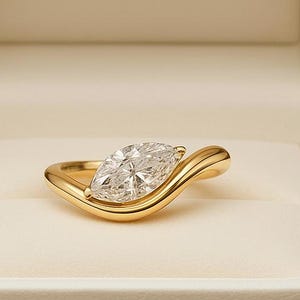Op de afbeelding: Een marquise-geslepen diamanten ring in een gouden zetting, gepresenteerd in een witte ringdoos. De ring heeft een grote, heldere diamant en een gebogen band. De doos is open en onthult de ring tegen een crèmekleurig interieur. De tekst "LUSTRELEGACY" is binnenin gedrukt.