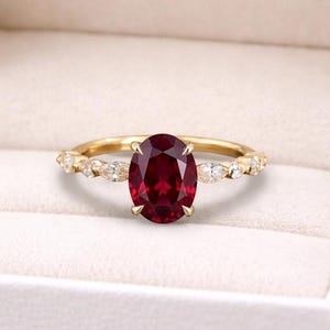 Pode incluir: Um anel de noivado dourado com uma grande pedra preciosa oval, vermelho escuro. O anel está em uma caixa branca. A banda apresenta pequenas pedras de destaque transparentes. O texto "Lusterlegacy" está no fundo.