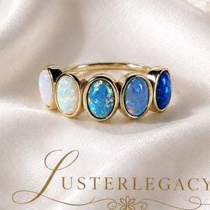 May include: A gold ring featuring five oval-shaped stones arranged in a row, transitioning in color from white to light blue and deep blue. The text LUSTERLEGACY is printed below the ring in a decorative script font with a small floral icon.