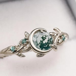 May include: A silver ring with a large, round, light green gemstone with dark green inclusions. The ring features a crescent moon design around the main stone and small blue-green accent stones. The band has a leaf and vine motif.