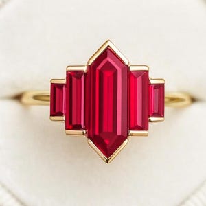 May include: A gold ring with a striking red gemstone, rectangular in shape, at the centre, with smaller rectangular stones on either side. The ring has a geometric design and is displayed in a white ring box.