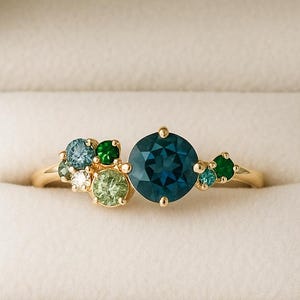 Op de afbeelding: Een gouden ring met een grote, ronde, diepblauwe edelsteen, omringd door kleinere, gekleurde edelstenen in groen- en blauwtinten. De ring wordt gepresenteerd in een witte sieradendoos.